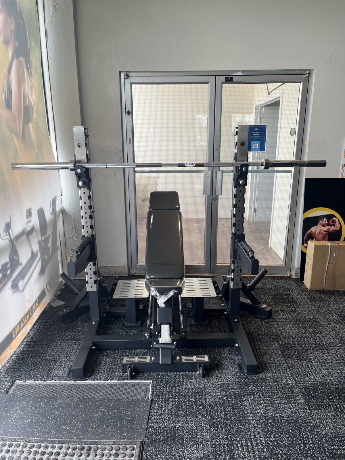 ARGO Fitness   Weightlifting bench and barbell on a squat rack in a small gym room with glass doors behind