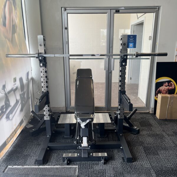 Weightlifting bench and barbell on a squat rack in a small gym room with glass doors behind.