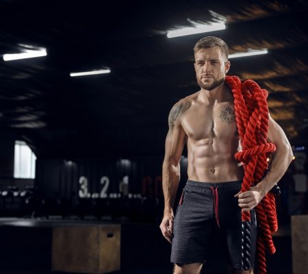 Young healthy male athlete doing exercises in the gym Young healthy man, athlete posing confident with the ropes in gym. Single male model practicing hard and training his upper body. Concept of healthy lifestyle, sport, fitness, bodybuilding, wellbeing.
